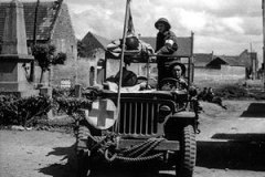 Le soldat L.E McKeating, conducteur d une Jeep ambulance, et le soldat L.F. McCadam, 9 C.I.B du 23ème régiment des ambulances de campagne, transportent deux brancards avec des blessés.27 juin 1944 BASLY